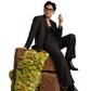 A man in a black suit and glasses sits on a large chocolate bar decorated with green icing and small green candies, holding a small gold card against a plain white background.