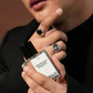 A close-up of a person holding a clear glass perfume bottle labeled 'PISTACHIO ANAFEK' with a black cap. The person is wearing a black turtleneck and silver rings, with a warm brown background.